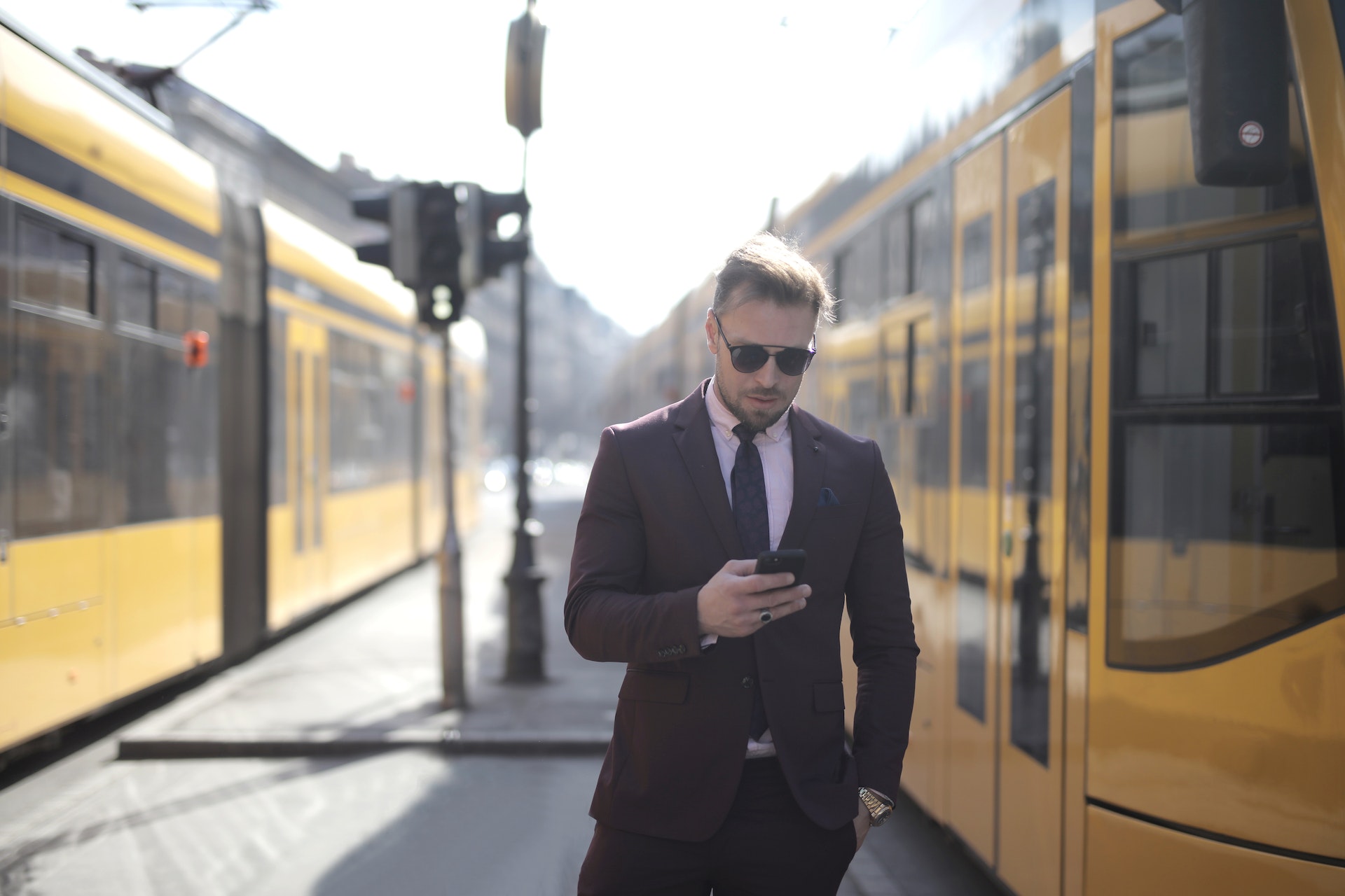 Brutal male entrepreneur in elegant suit and sunglasses standing with hand in pocket on street between trams and messaging on cellphone